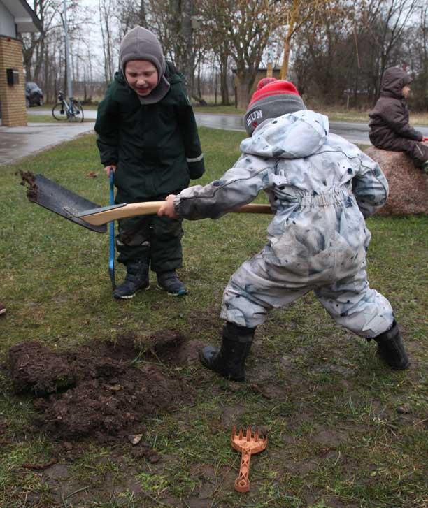 En dreng i flyverdragt har gravet et 10 cm dybt hul i græsplænen med en voksenskovl, mens der ved siden af ham står et andet barn og hepper.
