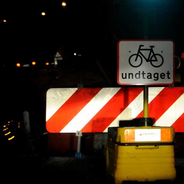 Mørk vejbane spærret af bom og skilt om undtagelse for cyklister. Skiltene oplyses af lyset fra forlygterne af en bil