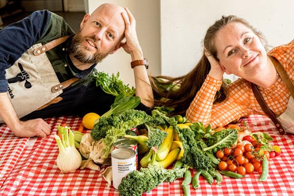 En mand og en kvinde ligger på et bord med en rødternet dug på og holder hver sit hoved. Imellem dem ligger en bunke af alverdens grønt, frugt og andre madvarer.