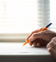 To rynkede hænder, den ene hånd holder et papir i en vindueskarm, den anden holder en kuglepen og er ved at skrive på papiret.
