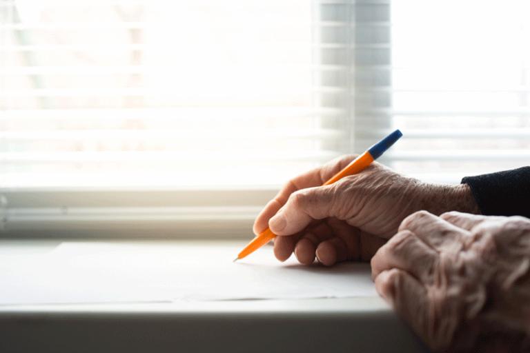 To rynkede hænder, den ene hånd holder et papir i en vindueskarm, den anden holder en kuglepen og er ved at skrive på papiret.