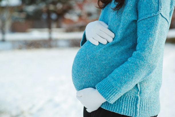 En gravid kvinde står i tyk trøje og holder med vanterne om sin mave. I baggrunden ligger der sne.