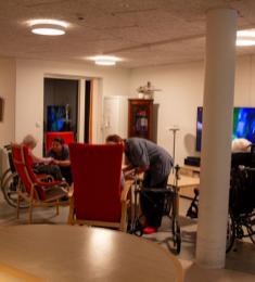 En dagligstue på et plejehjem, hvor tre ældre sidder foran fjernsynet, og to plejere i blå arbejdskitler er i dialog med de ældre.