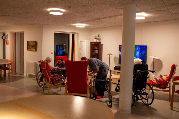 En dagligstue på et plejehjem, hvor tre ældre sidder foran fjernsynet, og to plejere i blå arbejdskitler er i dialog med de ældre.
