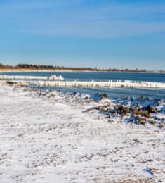 Kyststrækning med sneklædt strand med en bådebro, hvor der hænger is på. I baggrunden blå himmel.
