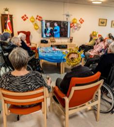 En gruppe af ældre sidder i bløde stole og kørestole foran fjernsynet. Mange har gyldne kongekroner på, mens statsministeren udråber den nye konge på fjernsynsskærmen.