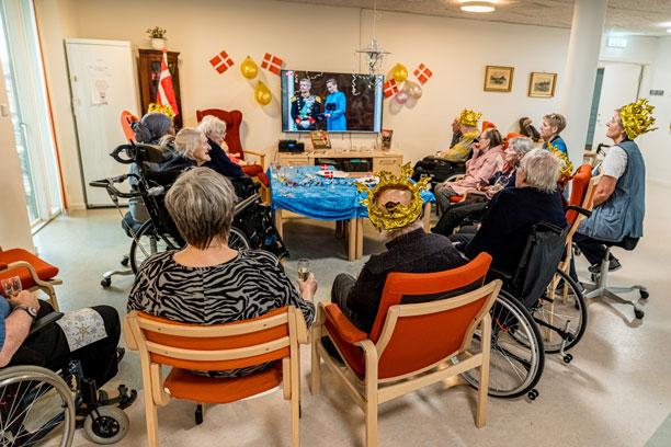 En gruppe af ældre sidder i bløde stole og kørestole foran fjernsynet. Mange har gyldne kongekroner på, mens statsministeren udråber den nye konge på fjernsynsskærmen.
