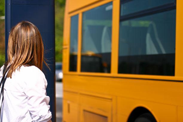 En kvinde står ved busstoppested, mens gul bus kører forbi hende.