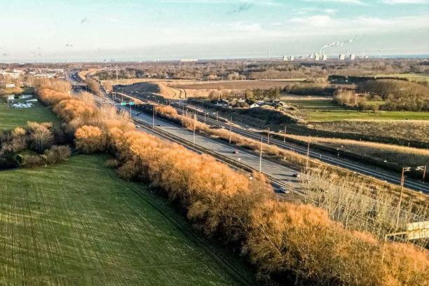 Dronebillede af fire spor med motorvej blandt grønne marker og gule og brune træer. I baggrunden Avedøre Værket
