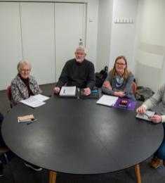 Fem mennesker sidder rundt om et rundt bord i et neutralt mødelokale. Alle har papirer eller notesblokke foran sig.
