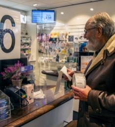 Ældre mand står ved skranken på et apotek med en bon og en rulle plastik i hånden.