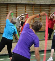 Ældre kvinder står i en gymnastiksal med ribber og spejle og laver gymnastik med en elastik.