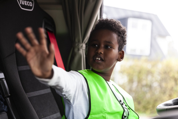 En dreng sidder på forsædet i en lastbil og vinker til nogen udenfor billedet.