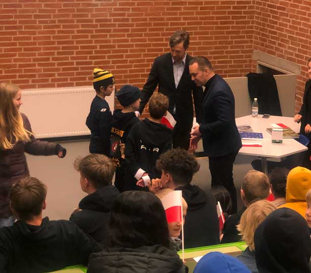 Tre drenge står foran alle deres skolekammerater i en større sal og taler med den polske ambassadør