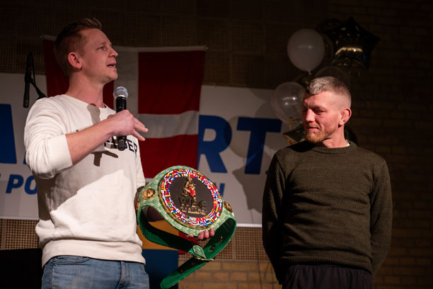 En mand med en mikrofon og et verdensmesterskabsbælte i hånden taler foran en anden mand på scenen, der kigger på bæltet..