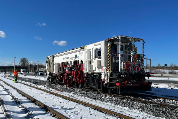 En stor skinnekørende maskine kører på et skinnestræk, hvor sneen ligger på begge sider af sporet.